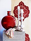 Decorative arrangement of red glass vase and white artistic candlesticks in front of ornamental wallpaper