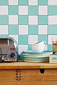 Stack of blue and yellow plates next to stainless steel toaster on wooden cabinet against turquoise and white chequered wall tiles