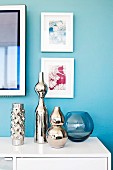 Mirrored vases of various sizes on white sideboard below framed pictures on wall painted pale blue