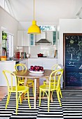 Yellow pendant lamp above round oak table and yellow-painted Thonet chairs on black and white striped rug in front of open-plan kitchen with drawings on blackboard to one side