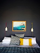 Verschiedene Kissen auf Doppelbett vor dunkelgrau gestrichener Ziegelwand mit Landschaftsbild und künstlerischen Wandleuchten