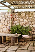 Vintage garden chair with cushion and rustic wooden bench on stone floor of terrace with bamboo pergola and tall stone wall