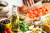 Olivenöl und Zutaten für ein Nudelgericht; im Hintergrund werden Tomaten kleingehackt
