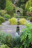Steinboden mit schmalen, eingelassenen Wasserrinnen und Ziergräser, im Hintergrund Wasserbecken vor traditionellem Pavillon in englischem Garten