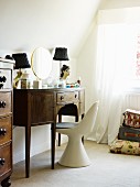 White shell chair in front of antique dressing table in dark wood next to window with curtain