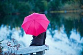 Frau mit pinkfarbenem Regenschirm am See sitzend
