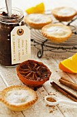 A jar of homemade mincemeat, mince pies on a rack, spices and half an orange