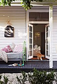 White wooden house, cat walking out through open French windows, comfortable vintage daybed with floral cushions on black wooden terrace