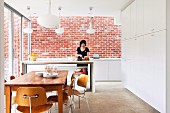 Dining area in front of kitchen counter, woman preparing food, exposed brick wall, tall white fitted cupboards and classic chairs in modern, open-plan kitchen