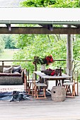 Rustikaler Holztisch und Stühle daneben Hund auf Tierfell liegend, im Hintergrund Rattan Sofa mit Kissen auf überdachter Holzterrasse vor sommerlicher Landschaft