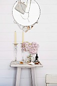 Glass candlesticks and vase of pink hydrangeas on white-painted console table below round mirror decorated with necklace and handbag on wooden wall
