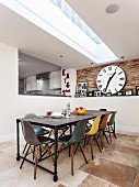 Long dining table, designer chairs of various colours, strip skylight, stone flagged floor and large wall clock on exposed brickwork