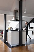 Kitchen area in converted attic, woman at white island counter on grey tiled floor behind black-painted wooden pillars