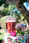 Floral tablecloth, refreshing drink in glass dispenser with tap and tray of glasses on garden table