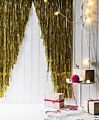 Glitzernder Fansenvorhang an weißer Bretterwand mit ausgeschnittener dreieckiger Tannenbaum Silhouettte, Beistelltisch mit Geschenken und Lichterkette