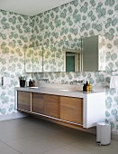 Long washstand with wooden sliding doors below mirrored wall units on wall with floral wallpaper