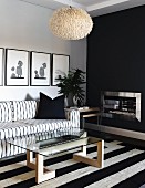 Coffee table with glass top on wooden base and black and white striped rug in front of black and white sofa below pictures on wall; open fireplace in black wall to one side