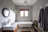 Free-standing vintage bathtub below window, modern washstand with wooden base cabinet opposite grey towels on hooks in bathroom painted pale grey