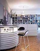 Bar stool at semicircular bar with illuminated glass top next to half-height, white chest of drawers used as partition in front of bookcase
