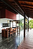 Frameless, sliding glass elements form transition between roofed wooden terrace and contemporary, open-plan kitchen with integrated dining area