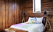 Simple double bed flanked by standard lamps with adjustable wooden bases in bedroom with dark wooden walls