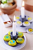 Simple candle stick and bottle caps painted yellow with various motifs arranged on white coaster