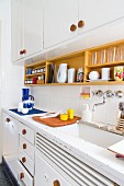 Küche in 60er Jahre Stil, Küchenzeile mit weisslackierten Hänge- und Unterschränken, an Türen und Schubladen Holzgriffe,über weisslackierter Arbeitsplatte Wandbord aus Holz mit Geschirr