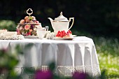 Romantisch gedeckter Gartentisch mit weißer Tischdecke, Goldrand-Kaffeeservice und üppiger Gebäck-Etagere mit Blütenschmuck