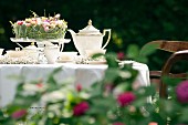 Romantisch elegant gedeckter Kaffee-Gartentisch mit weißer Tischdecke und Blumendekoration auf Glas-Tortenplatte