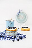 Tartlets with blueberries and cream, one in an enamel pan and one on a checked apron