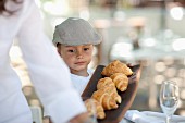 A little boy wearing a cap and holding a tray of fresh croissants