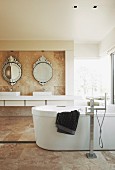 Free-standing bathtub in marble-tiled bathroom; washstand below round, ornate, antique-style mirrors