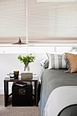 Modern bedside table next to bed with grey bed linen below window with closed roller blinds