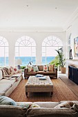 Helle Sofagarnitur und gepolsterter Couchtisch in mediterranem Wohnzimmer mit Blick durch Rundbogen Fenstertüren auf das Meer