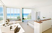 Half-height cabinet in front of bed and armchair next to panoramic window with sea view in spacious, white, light-flooded bedroom