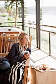 Woman sitting on pale wooden bench next to wooden stool on sunny balcony