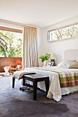 Bedroom bench at foot of double bed in bright, pleasant bedroom with blue-grey carpet and open glass wall leading to balcony