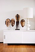 African masks on transparent plastic stand next to white table lamp on designer sideboard