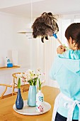Woman using feather duster to clean pendant lamp above vases of spring flowers on table