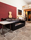 Wohn- und Arbeitsbereich mit Streichinstrumenten, rustikalem Steinboden, schwarzem Ledersofa und dunkelroter Wandfläche