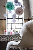 Feminine, romantic party decorations; pompoms, nostalgic lace tablecloth and flower arrangements on dining table in high-ceilinged, disused factory with industrial windows