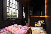 Schwarz gestrichenes Schlafzimmer mit Sprossenfenster, Bett mit bunter Tagesdecke und Retropolsterstuhl auf naturbelassenem Dielenboden