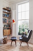 Cosy reading corner next to large lattice window, grey, retro wing-back chair with matching footstool, orange standard lamp and bookcase in niche