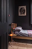 Blick in schwarz gestrichenes Schlafzimmer mit naturfarbenem Holzbett, Dielenboden und Tabletttisch mit beleuchteter Lampe und Rosensträußchen