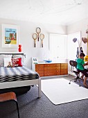 Child's bedroom - boy sitting on chair next to coat rack, bed, sideboard and tennis racquets hung on wall