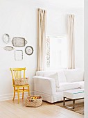 Korb auf Boden und gelb lackiertem Stuhl, an Wand Silber Tablettsammlung, daneben weisses Polstersofa vor Fenster mit bodenlangen Vorhängen