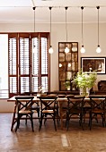 Long wooden table, bistro chairs and row of industrial-style pendant lamps; vintage decor with folding interior shutters and stone floor