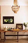 Rustic wooden table decorated with ceramics, china, vase of flowers and dish of fruit below framed still-life artworks and chandelier