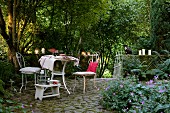 Atmospheric terrace seating area - white vintage metal chairs and table on cobbled floor in front of lit candles on low wall in densely planted garden