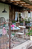 Gartenstühle und Tisch aus Metall auf Veranda, Zäune aus altem Metallgitter vor rustikalem Häuschen mit Mauerwerk und teilweise Holzverkleidung